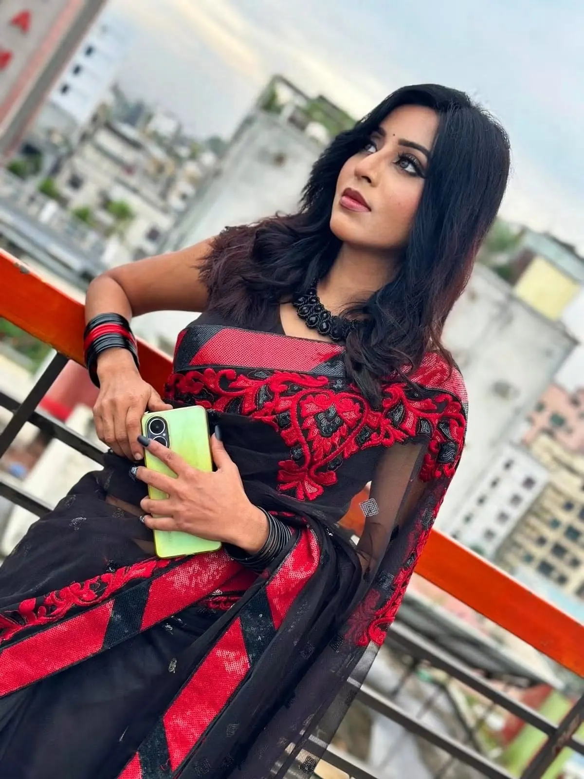 Sumona in a black and red saree holding a phone during an outdoor shoot